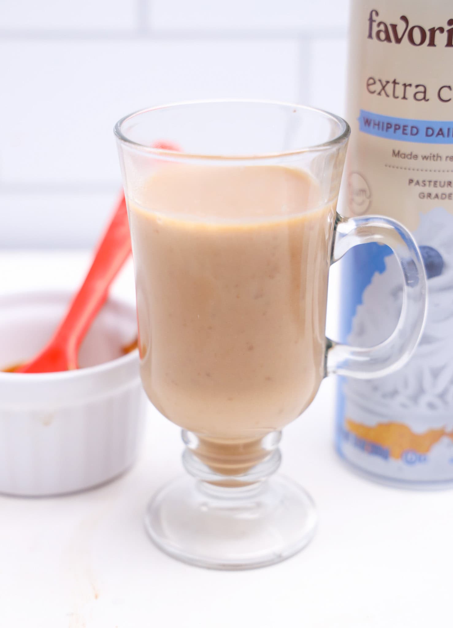 A glass mug of light brown Biscoff Hot Chocolate sits beside a white ramekin and a can of whipped dairy topping.