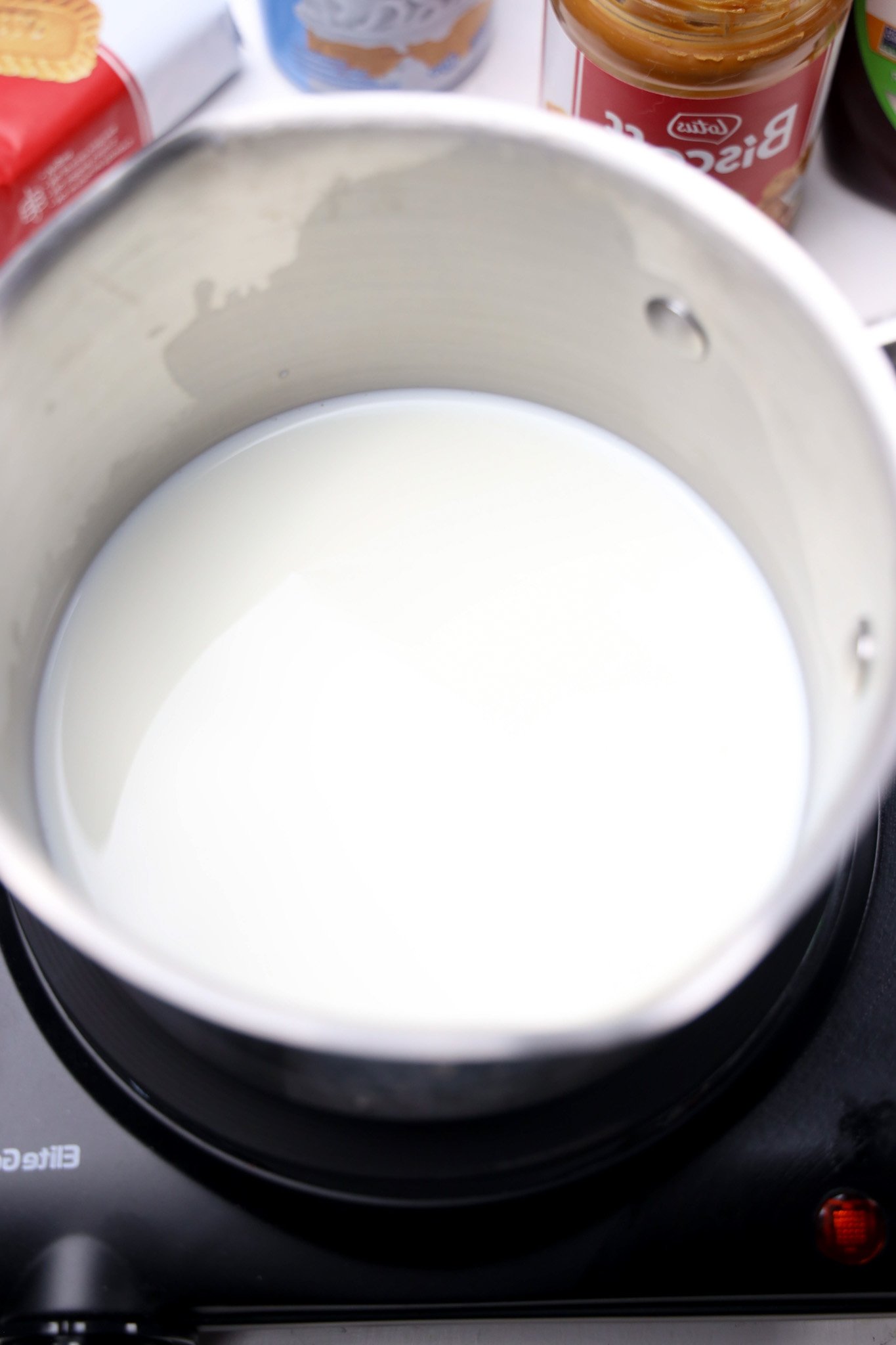 A pot of milk heating on a black electric stove, ready to be transformed into creamy Biscoff Hot Chocolate, with jars in the background.