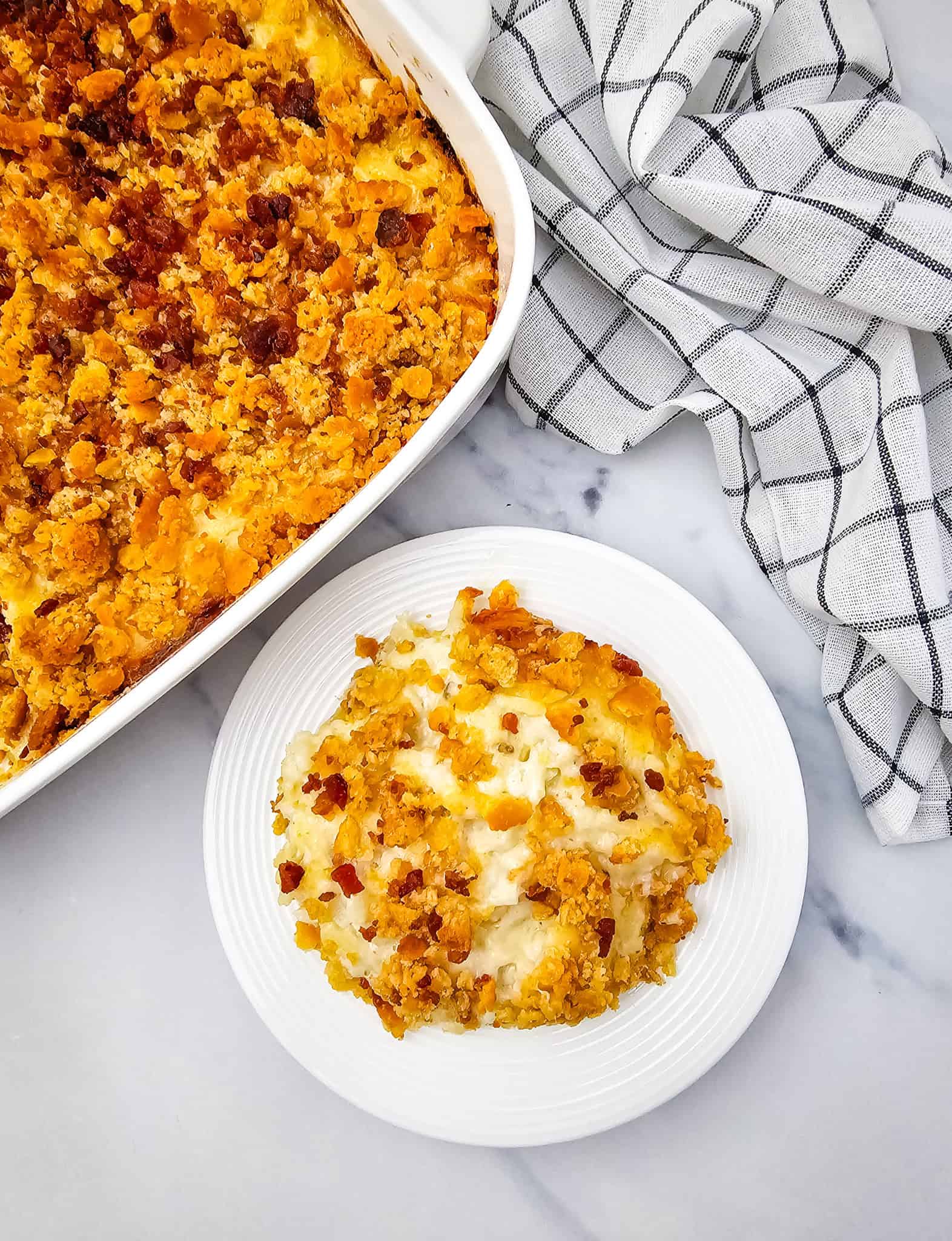 A serving of cheese potato casserole with a crispy topping sits on a white plate next to the casserole dish, showcasing layers of gooey cheese perfection.