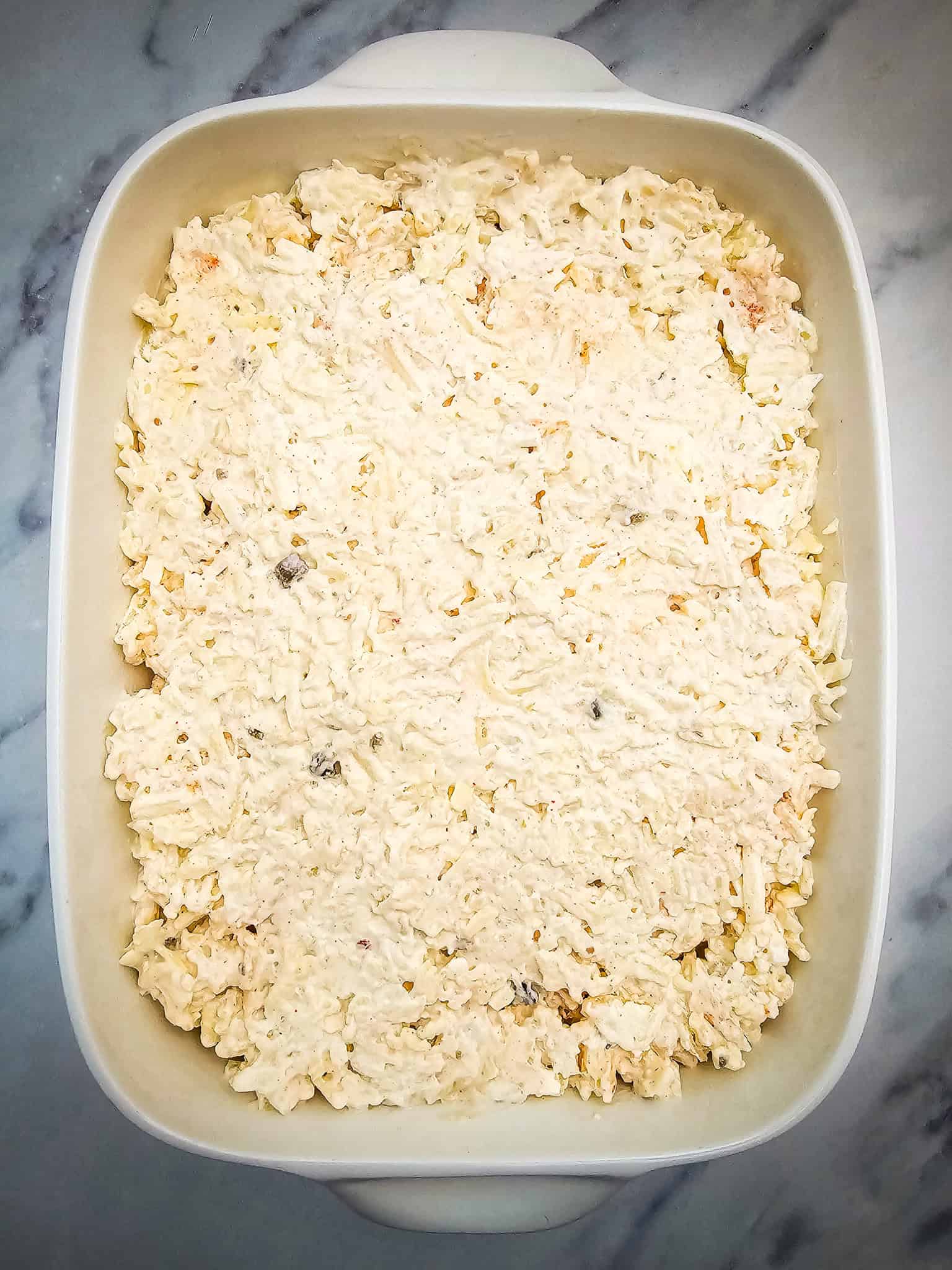 A white casserole dish filled with an uncooked creamy cheese and hashbrown mixture sits on a marble surface.