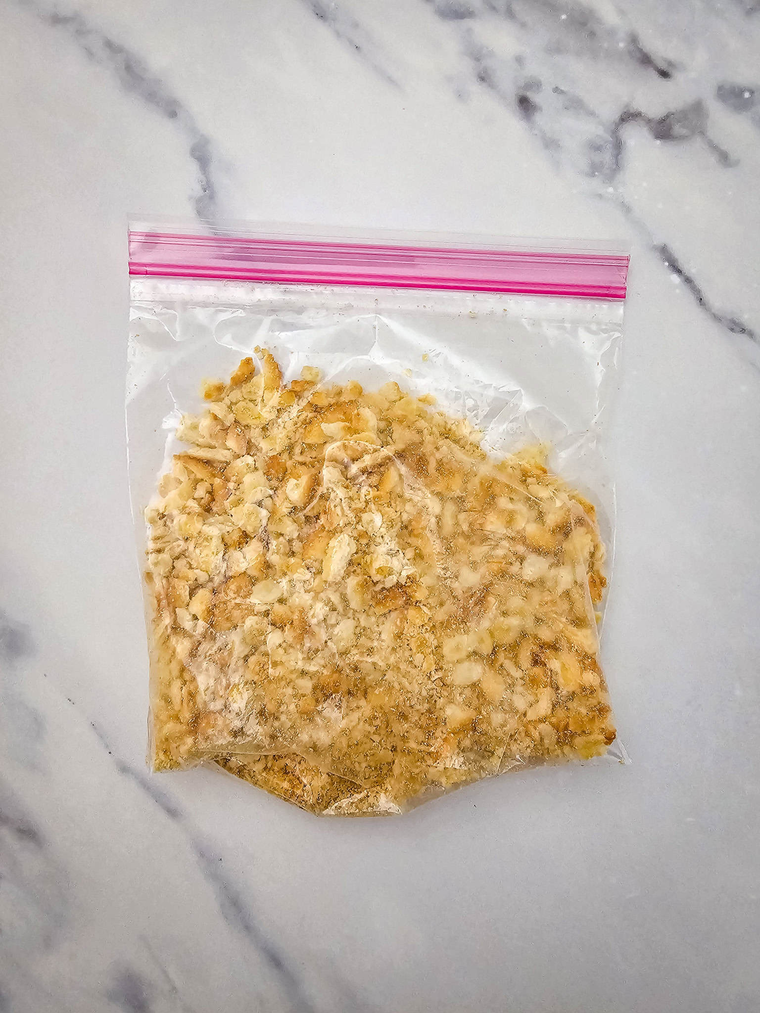 A plastic bag with crushed crackers, perfect for topping a cheesy potato casserole, sits on a white marble surface.