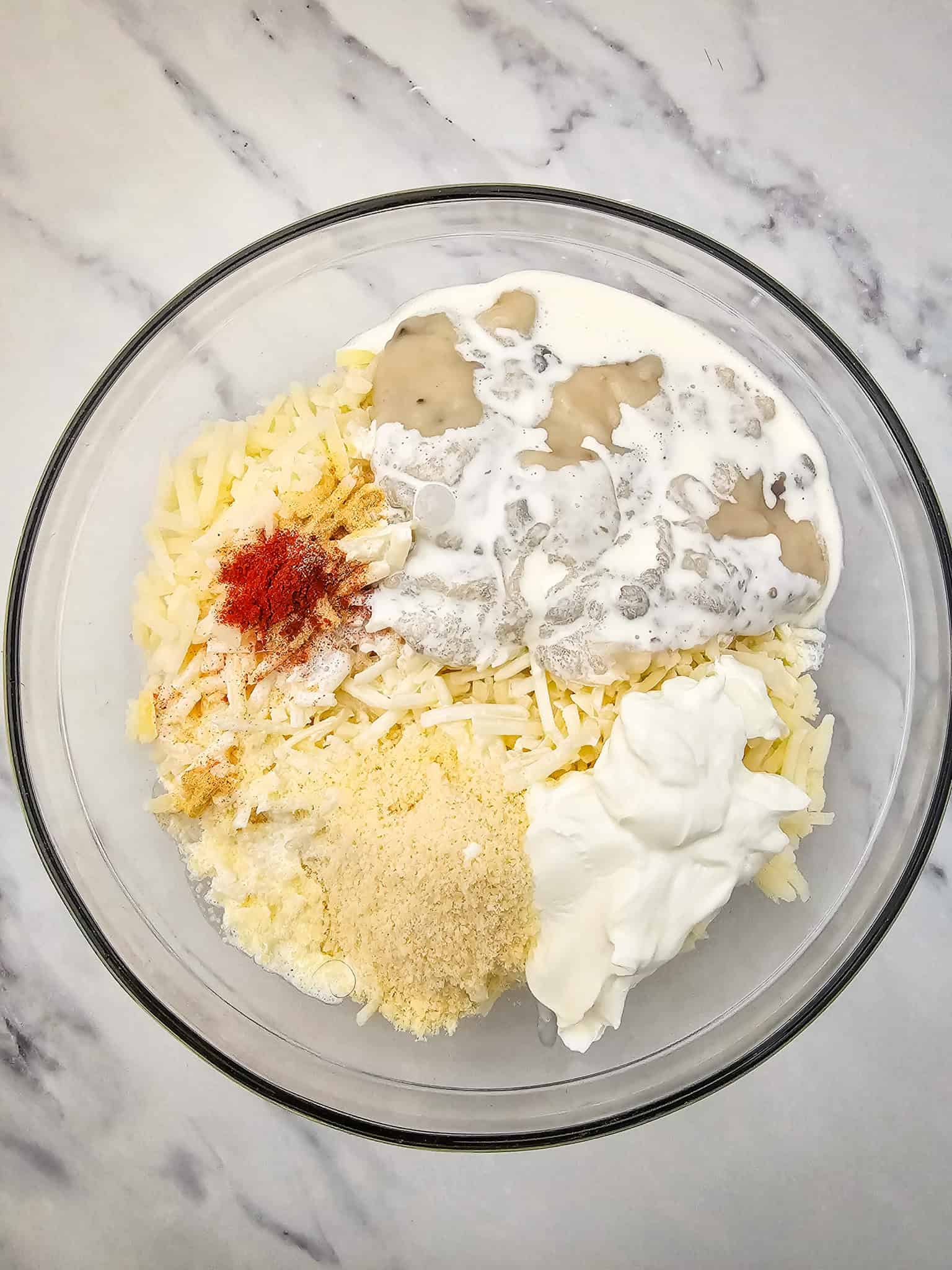 A glass bowl with shredded cheese, sour cream, cream, breadcrumbs, spices, and potato on a marble surface-perfect ingredients for a cheesy potato casserole.