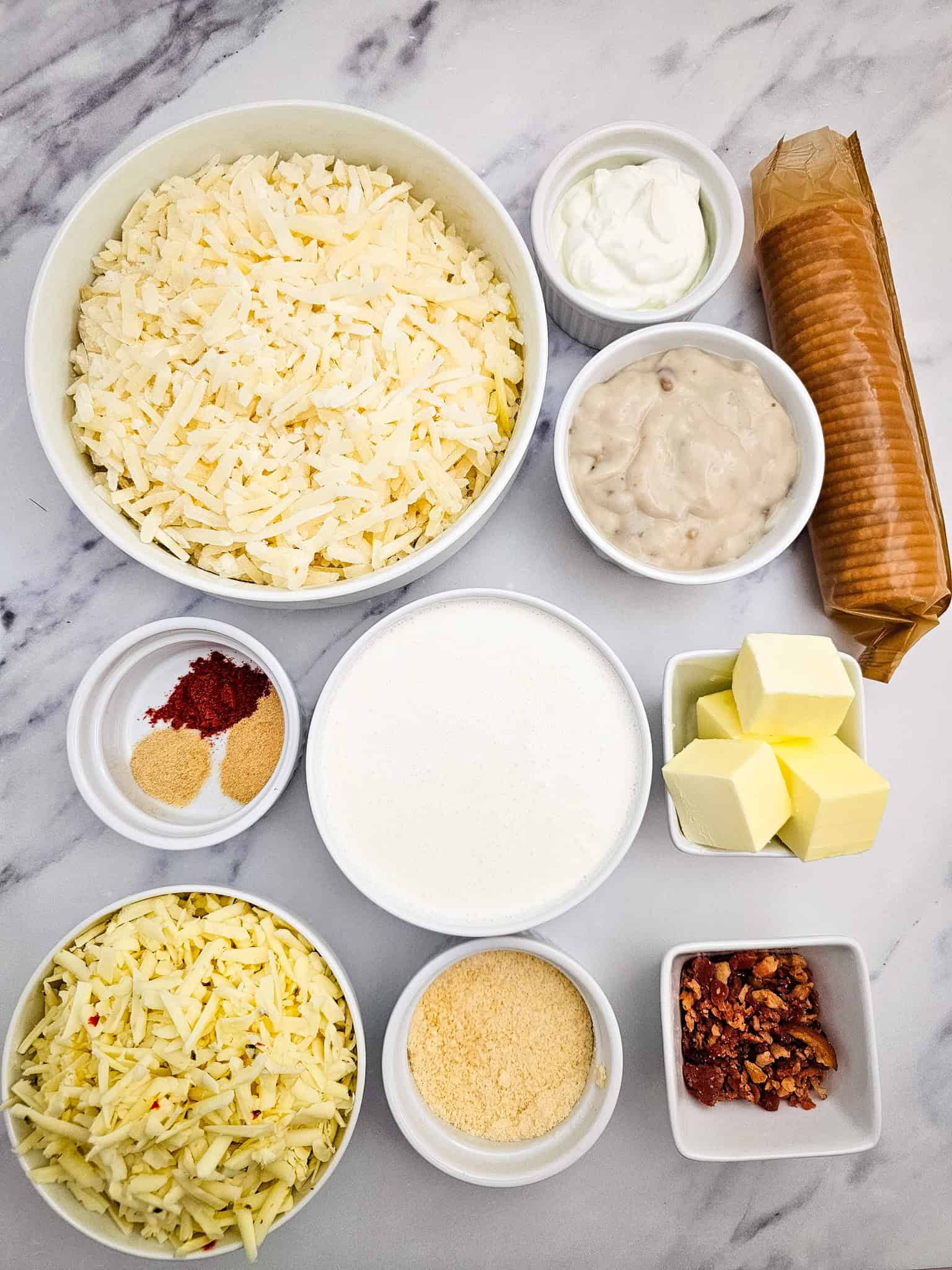 Bowls of shredded potato, cheese, butter, cream, seasonings, crackers, bacon, and sour cream on marble—perfect for assembling a comforting casserole.