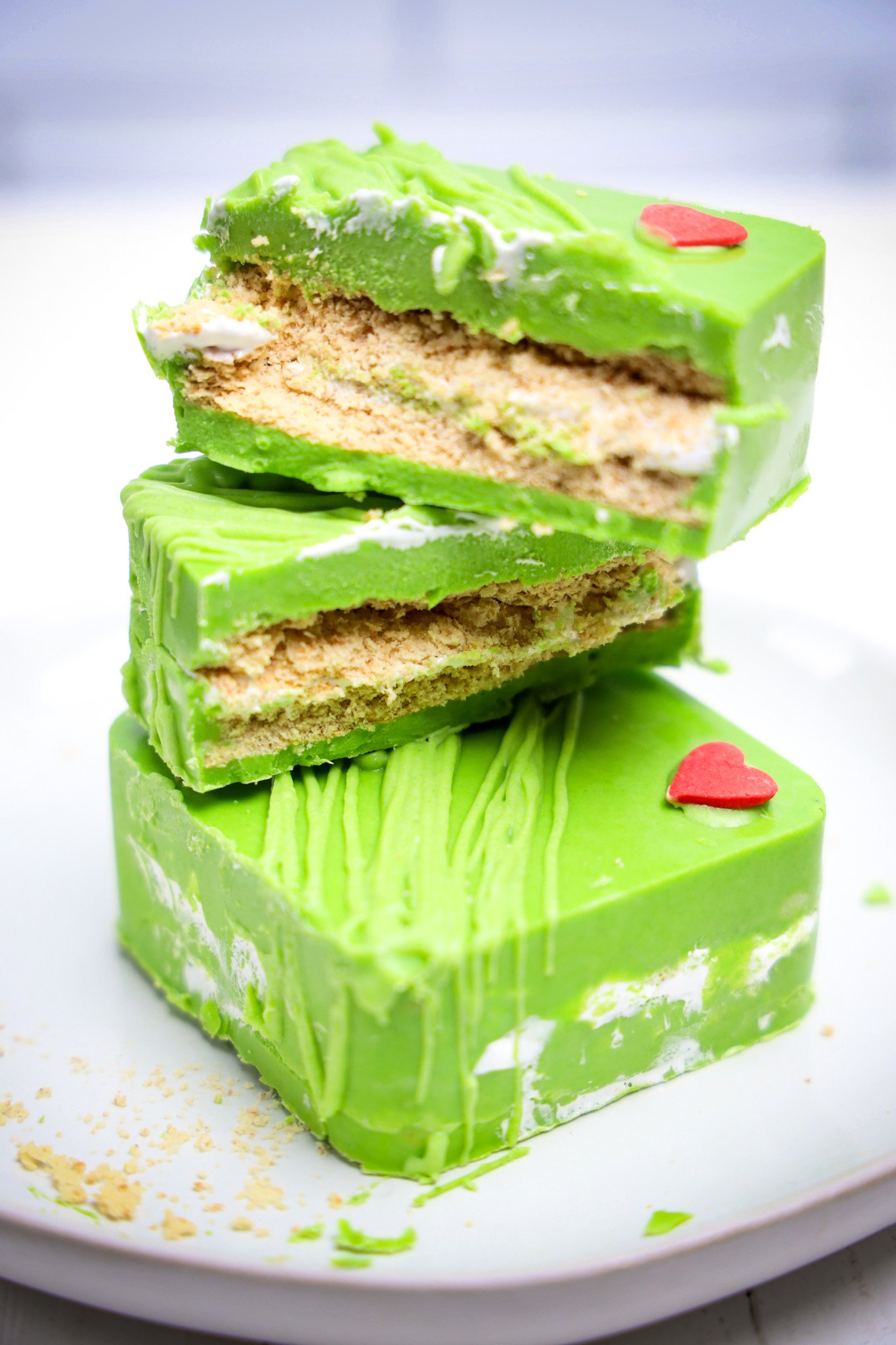 Three green s'mores sandwiches stacked, each with a red heart decoration, on a white plate—a festive Grinch dessert perfect for any holiday sweets table.