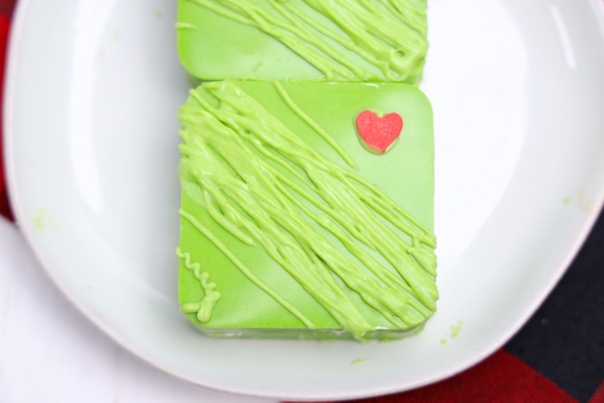 A green frosted Grinch dessert, shaped in a square with a red heart decoration, sits on a white plate.