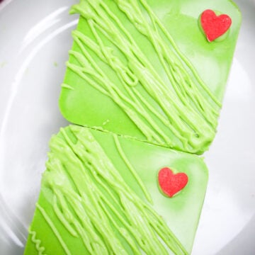 Two square green frosted cookies with red heart decorations on a white plate, inspired by a festive Grinch dessert, perfect for anyone craving fun and colorful Grinch S'mores Bars.