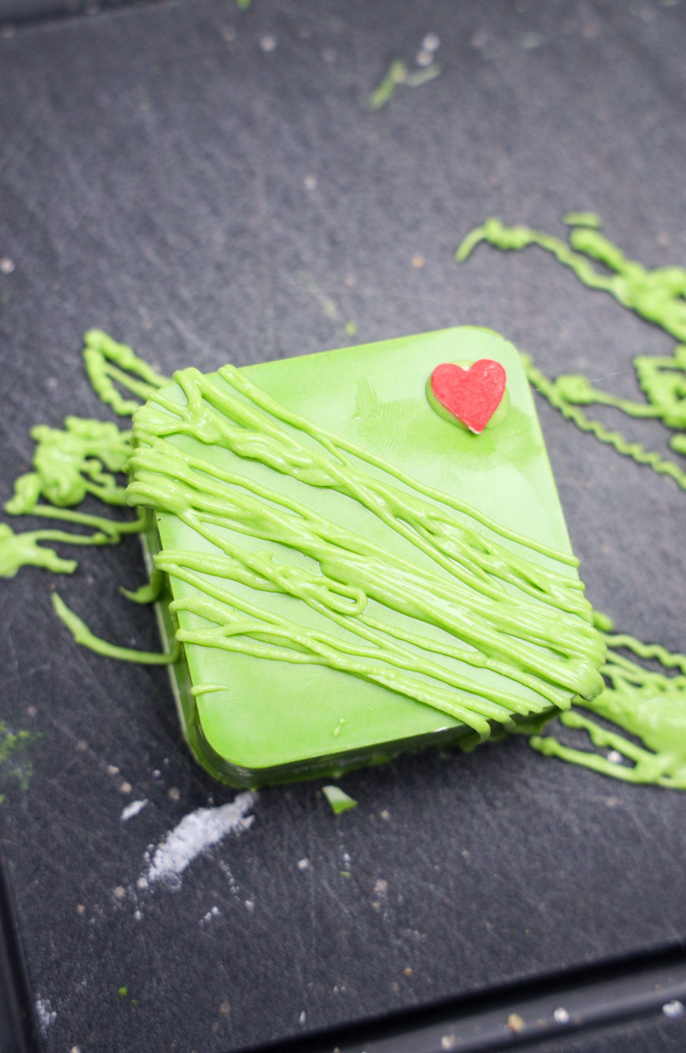 A Grinch S'mores Bars dessert: a green square treat with drizzled icing and a small red heart on top, placed on a dark surface.