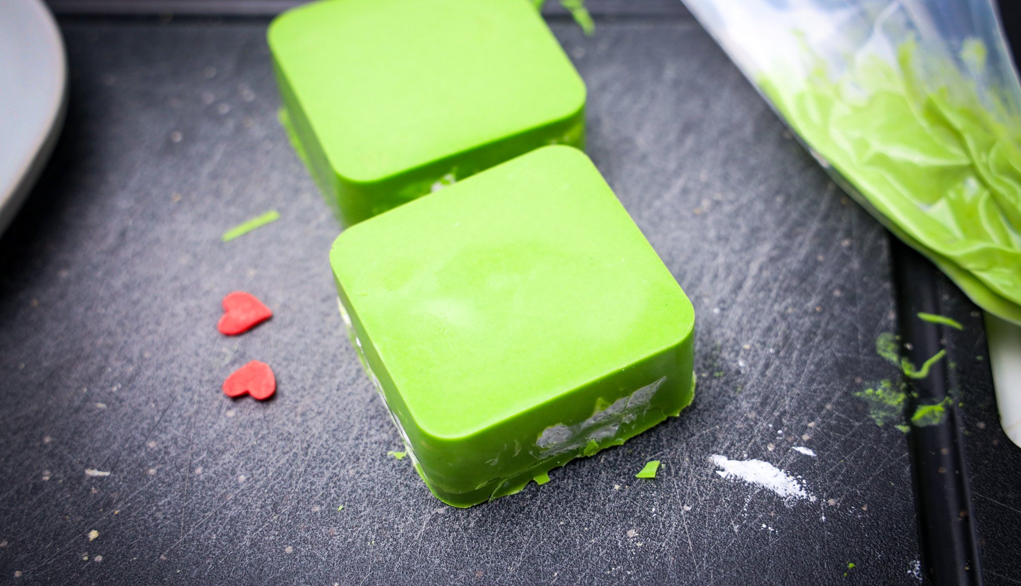 Two bright green, square-shaped Grinch S'mores Bars sit on a black surface, with two small red heart sprinkles nearby.