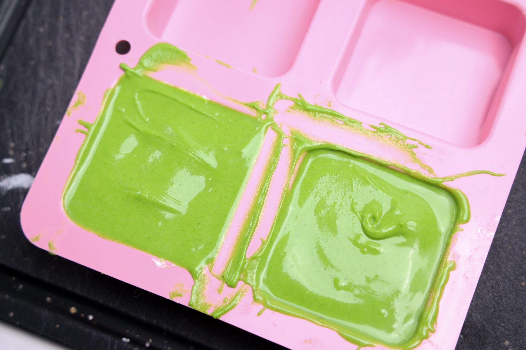 Green melted candy coating for Grinch S'mores Bars is spread in a pink silicone mold with two square compartments, resting on a black surface.