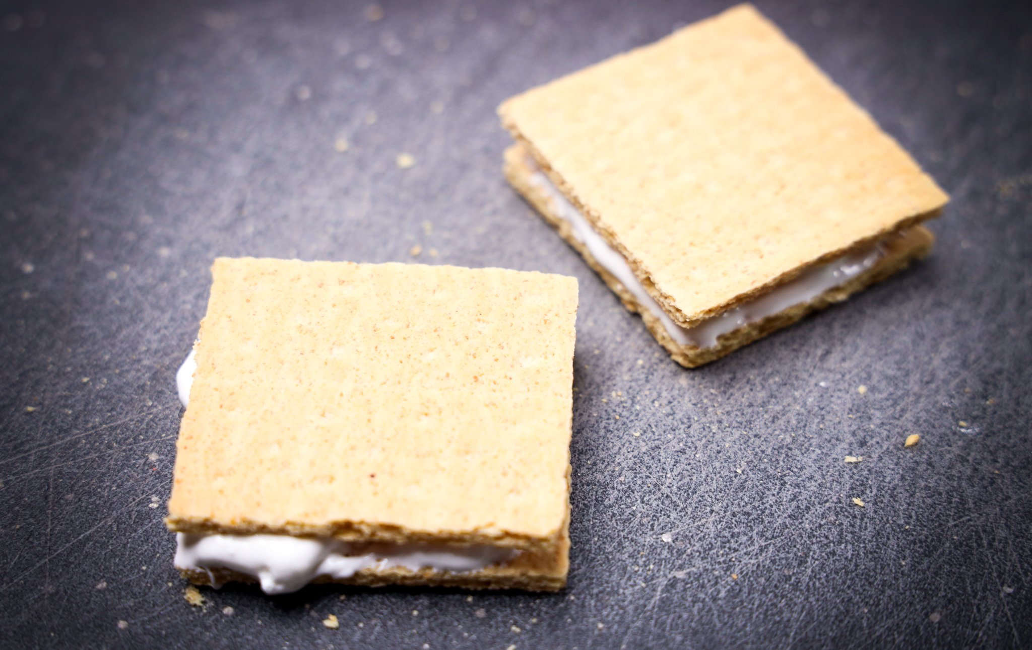 Two s'mores made with graham crackers and marshmallow filling on a dark surface