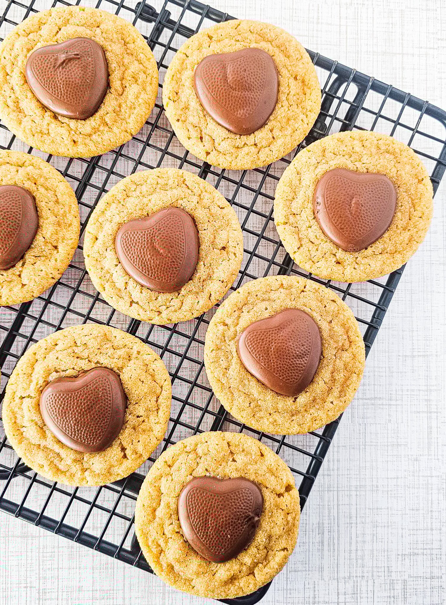Nine Easy Peanut Butter Blossoms topped with heart-shaped chocolates rest on a black cooling rack over a white background.