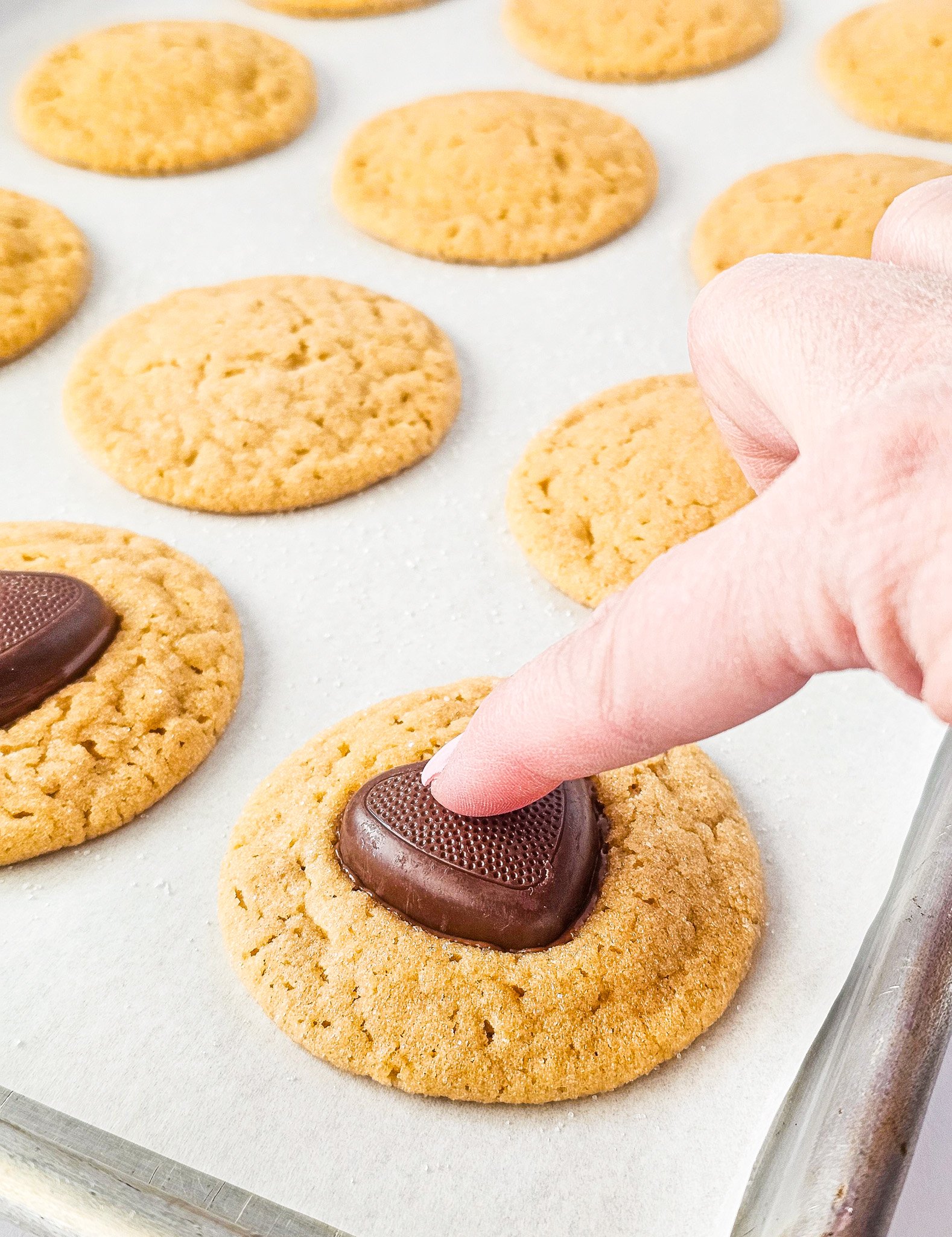 A hand presses a chocolate heart onto a freshly baked Easy Peanut Butter Cookie on a parchment-lined baking tray.