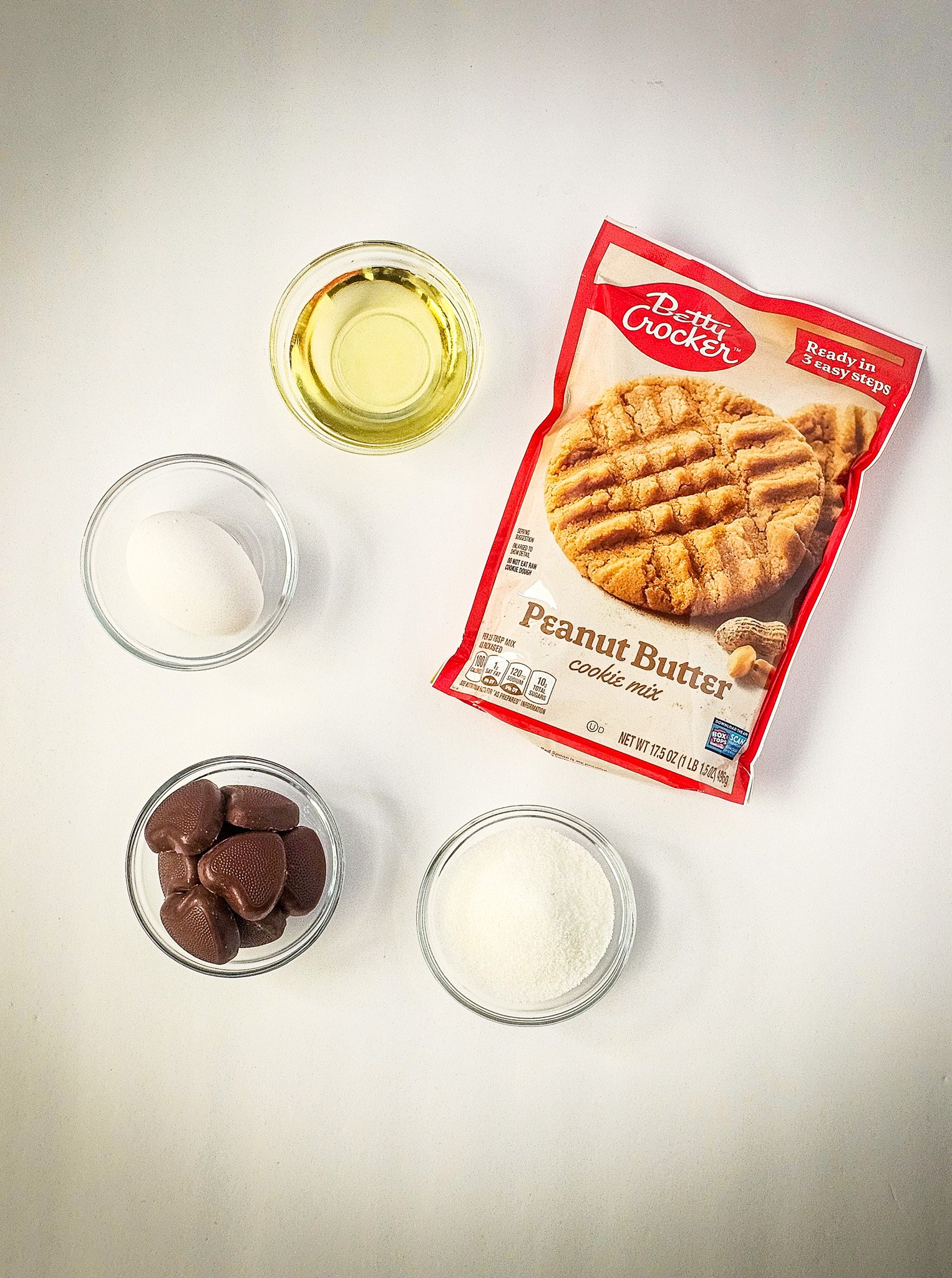 Betty Crocker peanut butter cookie mix, egg, oil, sugar, and chocolate in bowls on a white surface—everything you need for Easy Peanut Butter Cookies.