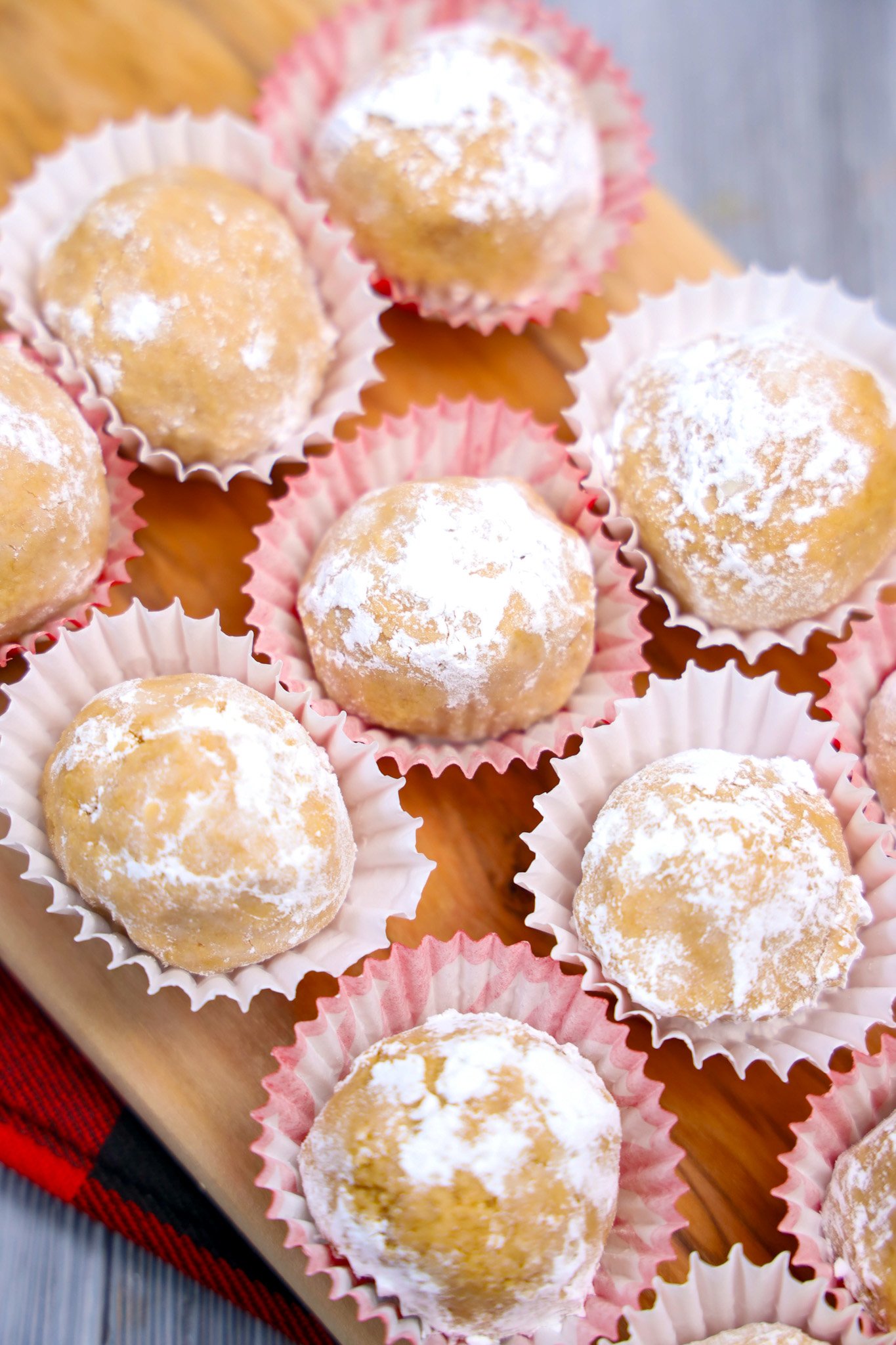 Peanut Butter Snowballs—powdered sugar-coated cookie balls with a rich peanut butter center—rest in paper liners on a wooden board.