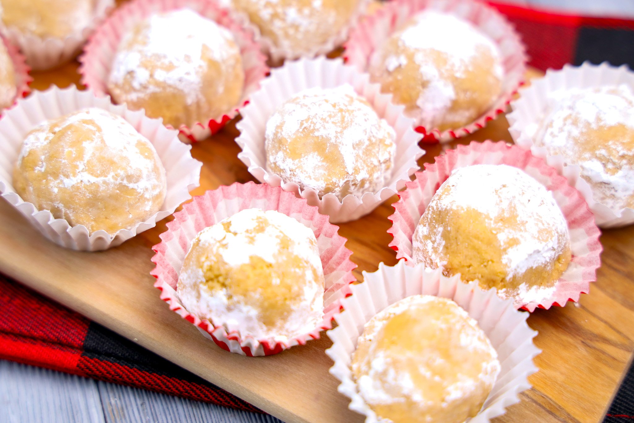Powdered sugar-covered Peanut Butter Snowballs in paper cups rest on a wooden board with a plaid cloth underneath.