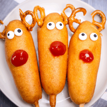 Three corn dogs decorated as Rudolph reindeer with pretzel antlers, candy eyes, and ketchup noses on a white plate.