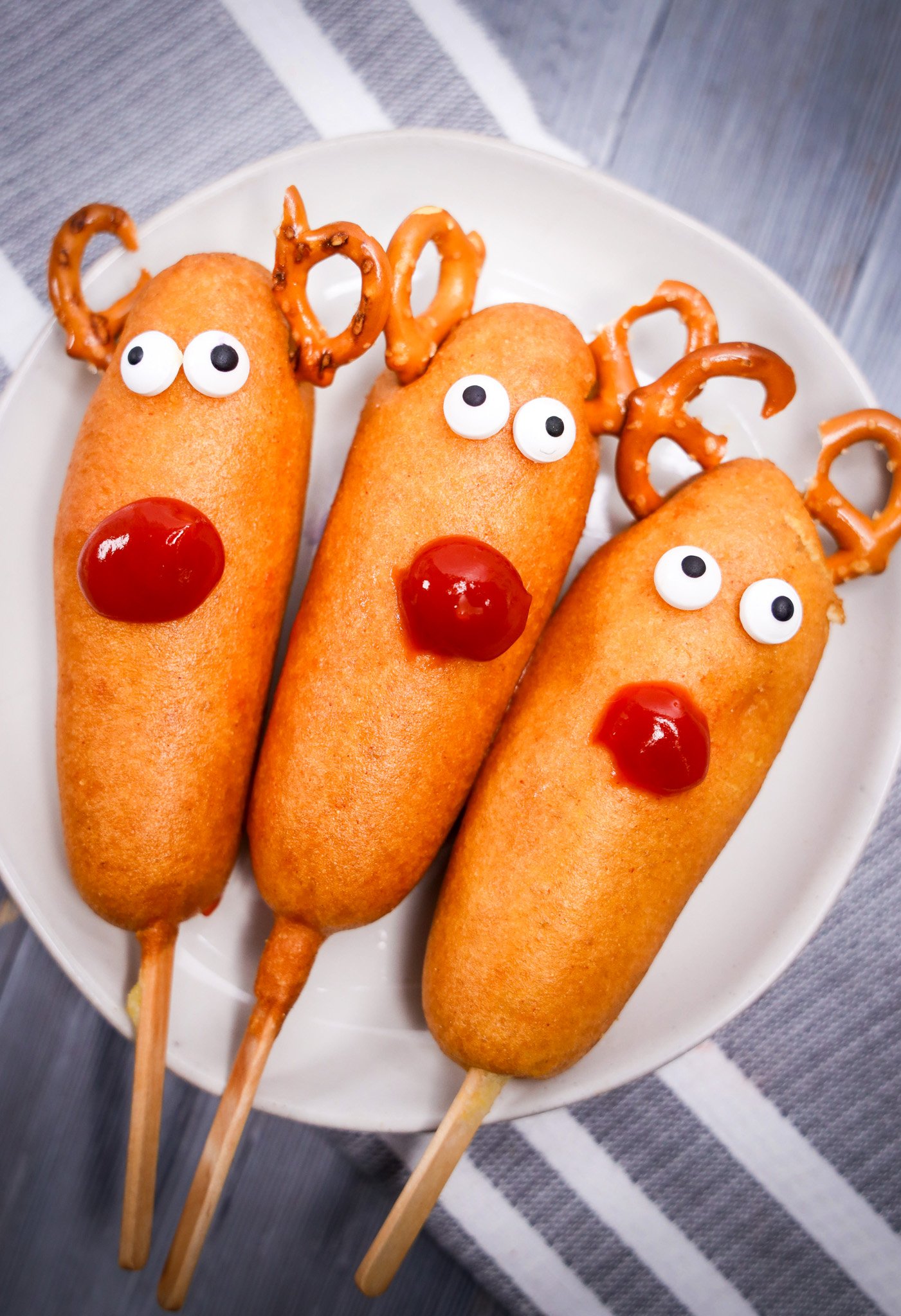 Three Rudolph Corn Dogs decorated as reindeer with pretzel antlers, candy eyes, and ketchup noses on a white plate.