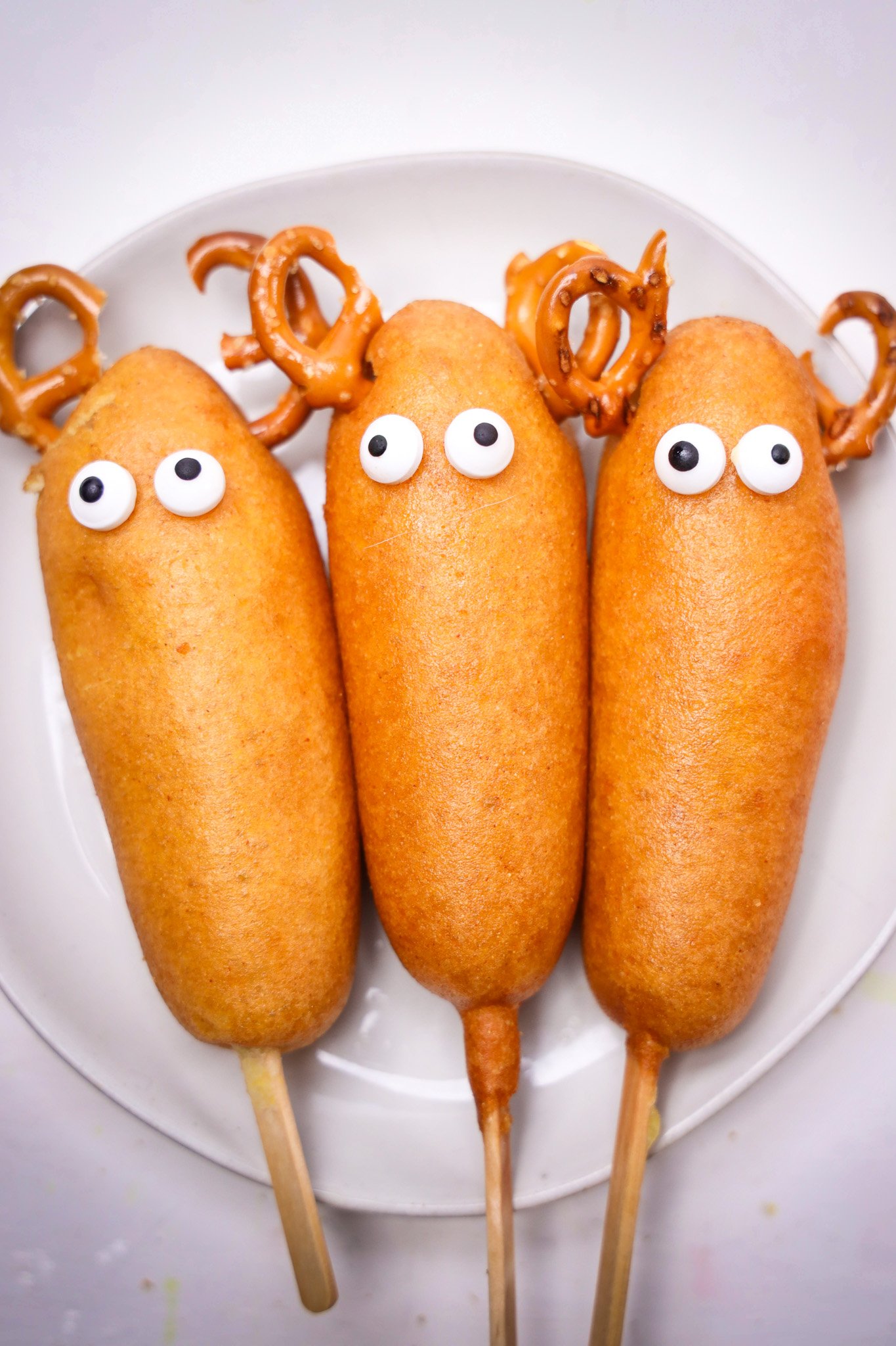Three corn dogs with candy eyes and pretzel antlers are arranged to look like Rudolph and his reindeer friends on a white plate.