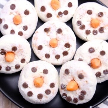 Powdered donuts are transformed into an easy Donut Snowman dessert, decorated with chocolate chips and orange candies as snowman faces on a black plate.