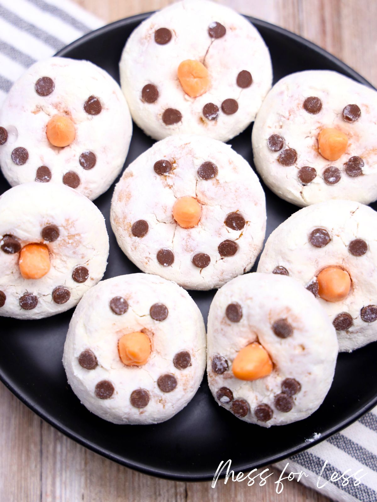 Powdered donuts are transformed into an easy Donut Snowman dessert, decorated with chocolate chips and orange candies as snowman faces on a black plate.