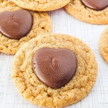 Easy Peanut Butter Cookies topped with heart-shaped chocolate pieces sit on a white textured surface, creating a delightful twist on classic Peanut Butter Blossoms.