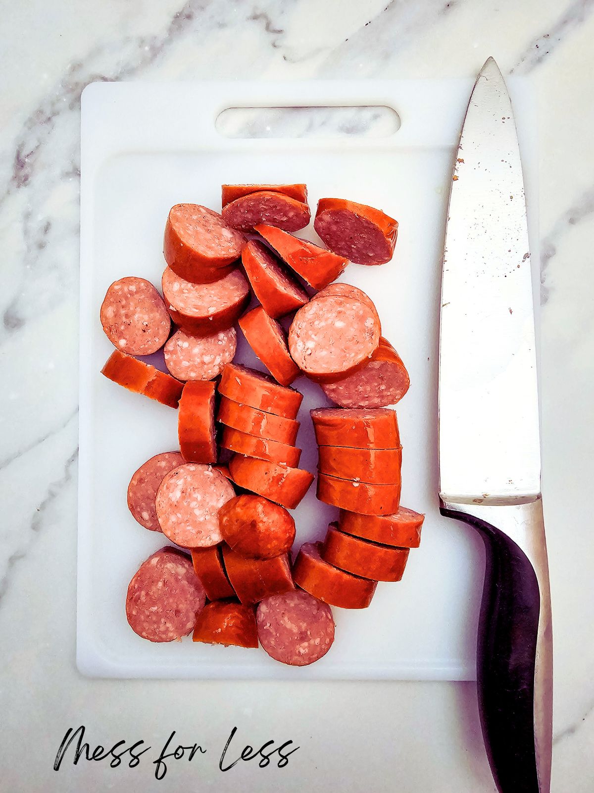 Sliced sausage and fresh veggies rest on a white cutting board with a knife, ready for your next sheet pan meal on a marble surface.