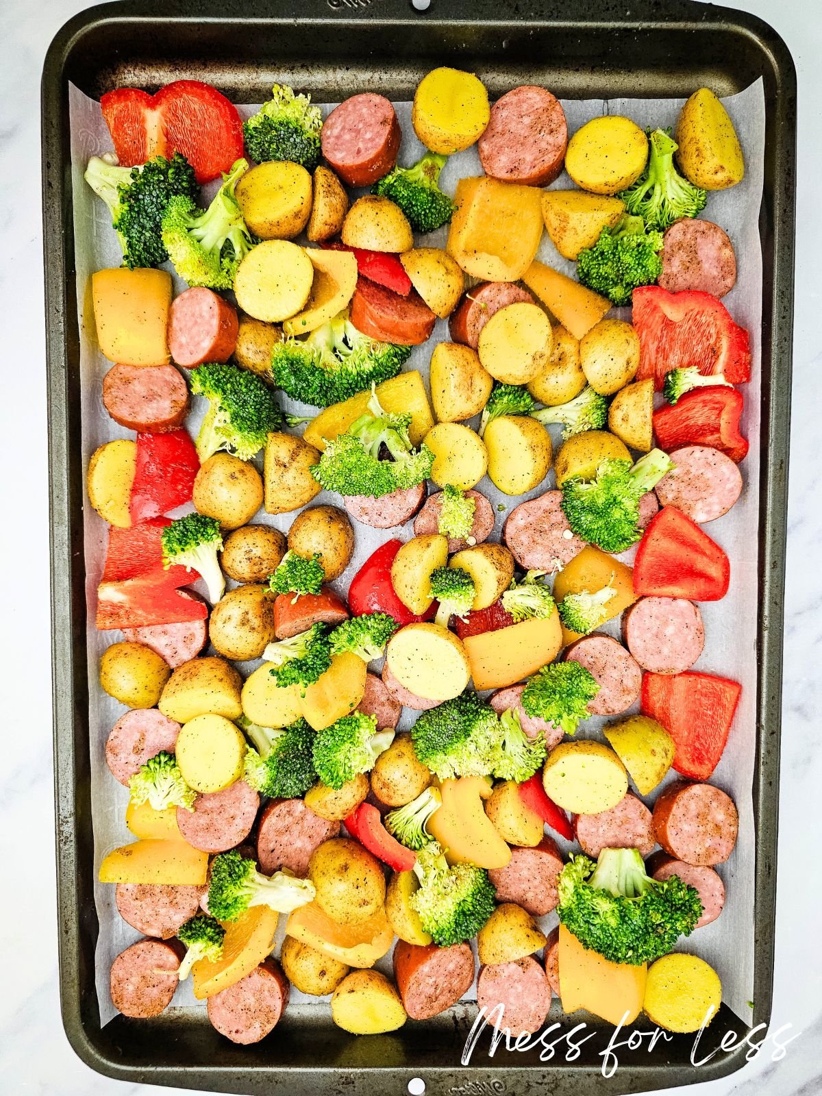 Sheet pan with sliced sausage, veggies like broccoli and red bell pepper, plus yellow potatoes, ready to be roasted.