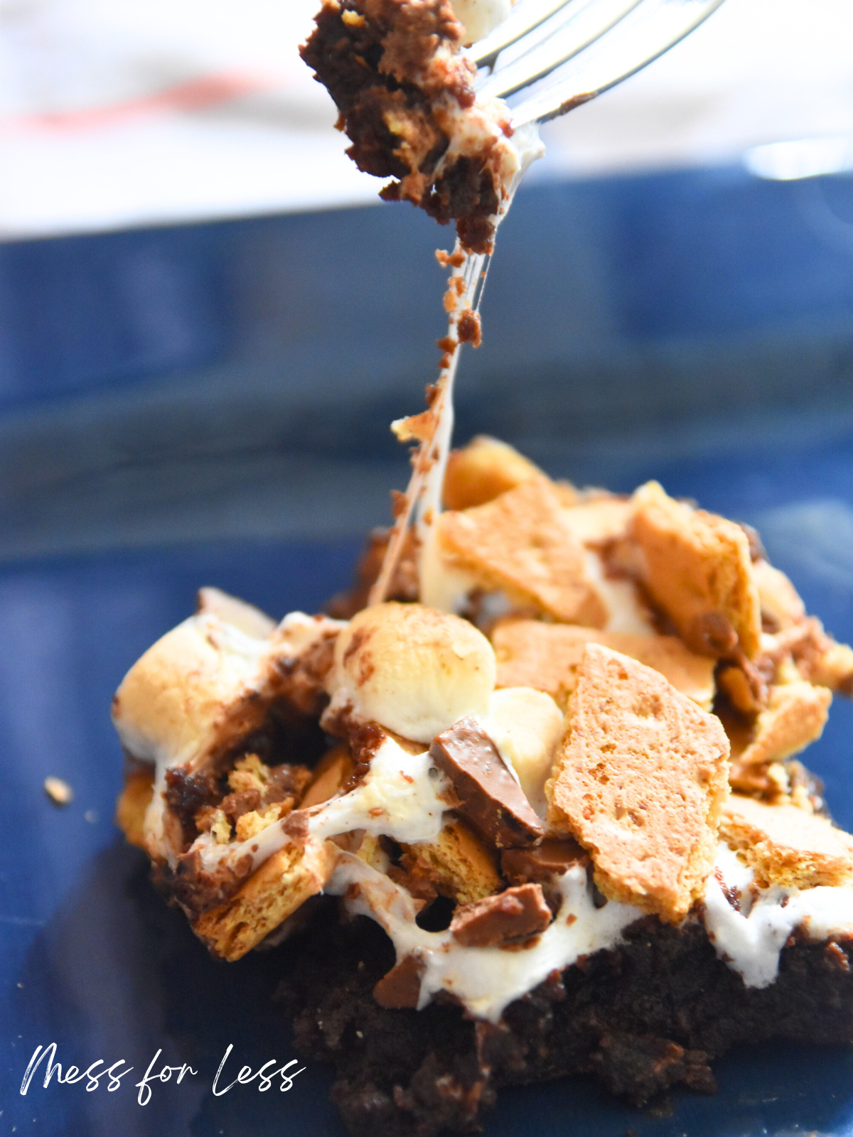 A gooey s’mores brownie made with boxed brownie mix is lifted by a fork, with melted marshmallow, chocolate, and graham cracker pieces.