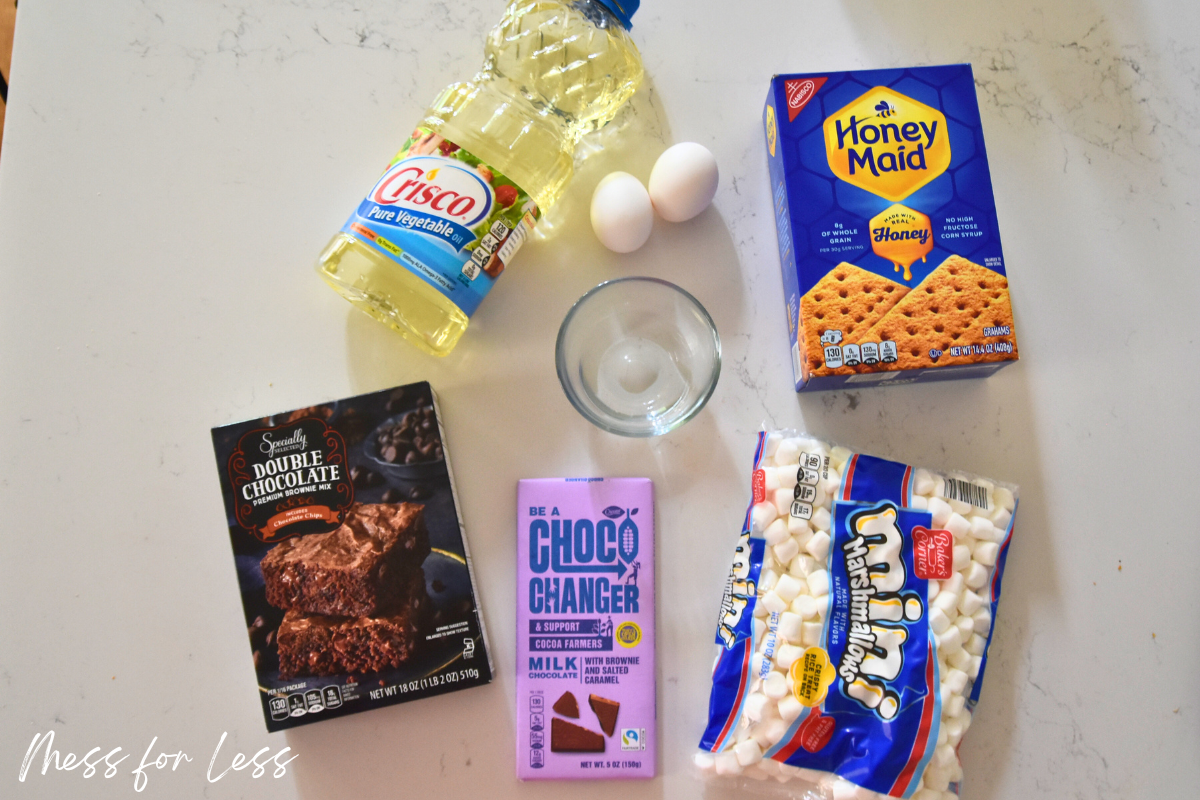 All the ingredients you need for S’mores Brownies—boxed brownie mix, a chocolate bar, marshmallows, graham crackers, eggs, oil, and a glass bowl—are laid out on the counter.