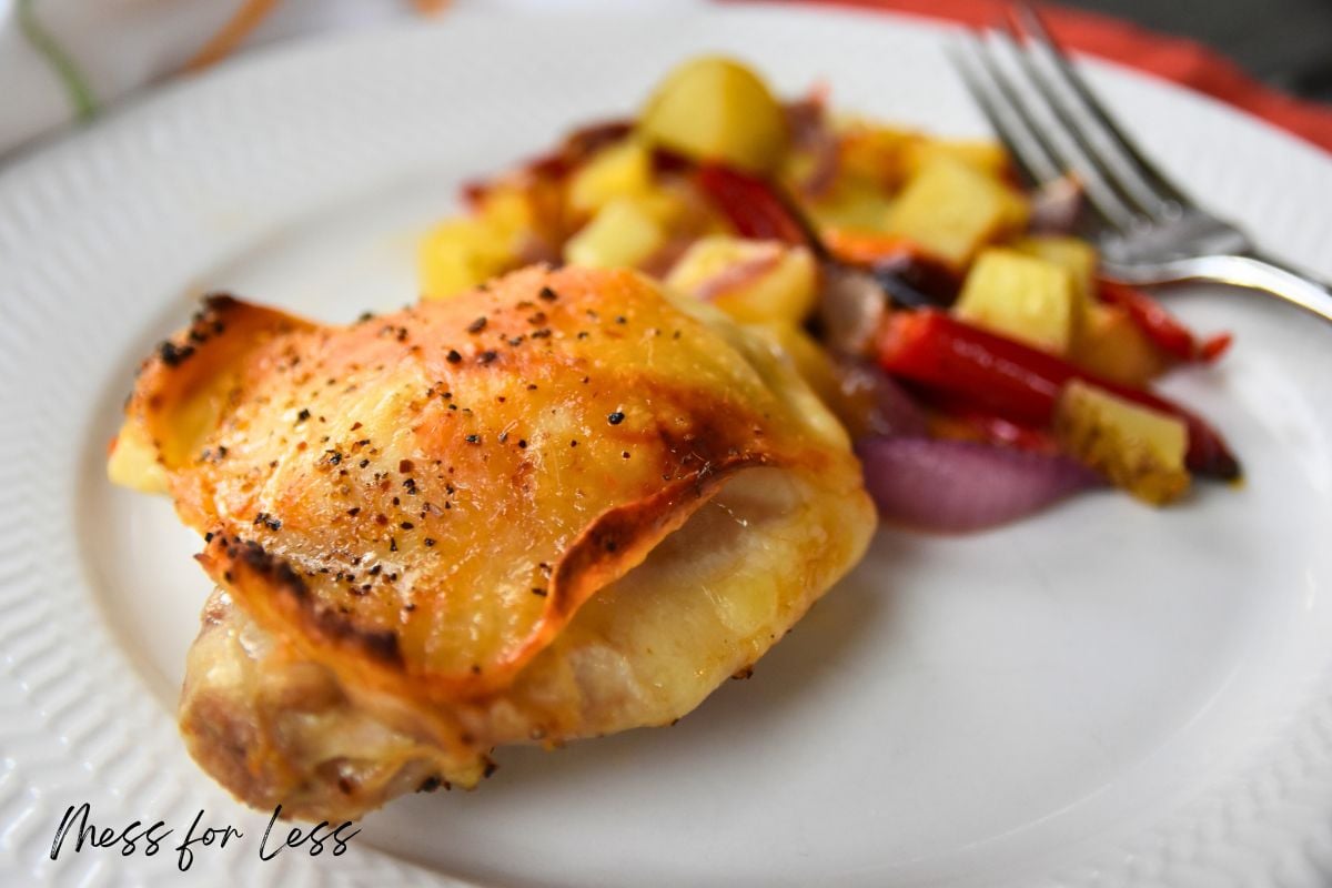 Baked chicken thigh with crispy skin, served with roasted potatoes, red onions, and red peppers—a delicious Sheet Pan Chicken Thighs recipe on a white plate.