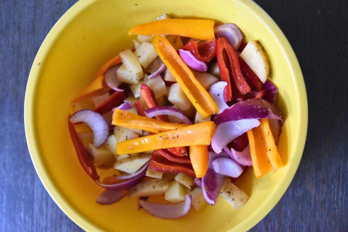 Sliced red onions, yellow peppers, and potatoes in a yellow bowl on a dark surface—perfect veggies to complement your next sheet pan chicken thighs recipe.